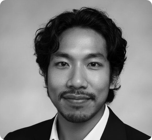 Black and white headshot of a young man with wavy hair, a mustache, and beard, wearing a suit jacket and white shirt, smiling slightly at the camera.