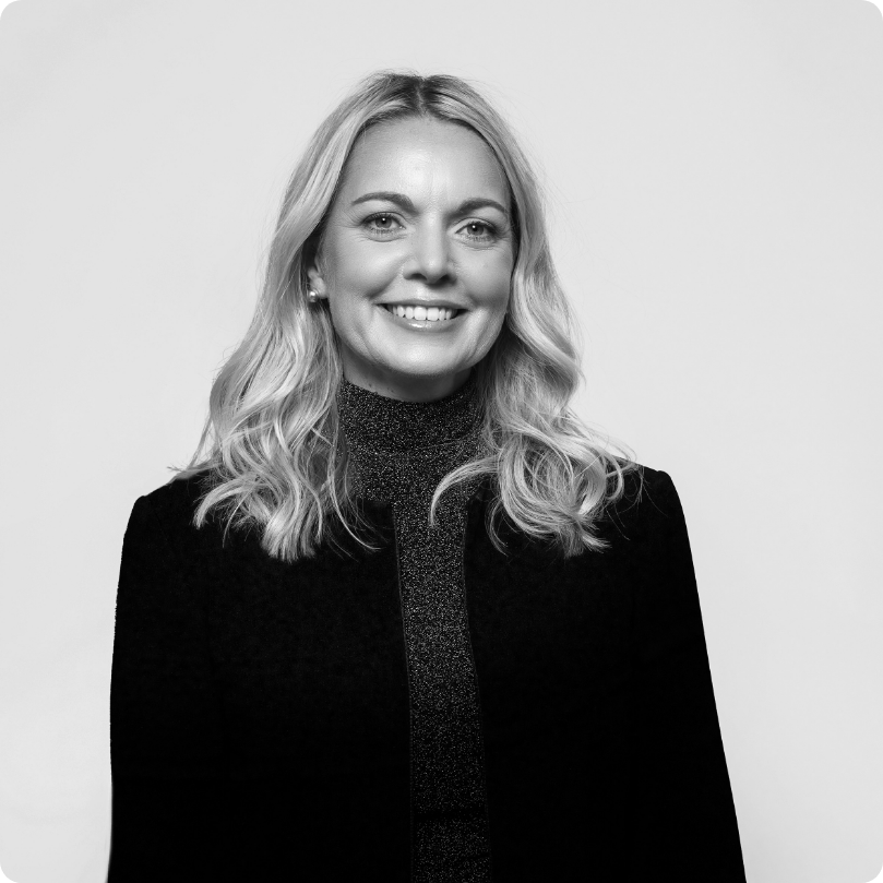 Black and white portrait of a smiling woman with blonde wavy hair, wearing a dark blazer and a glittery top, against a plain background.