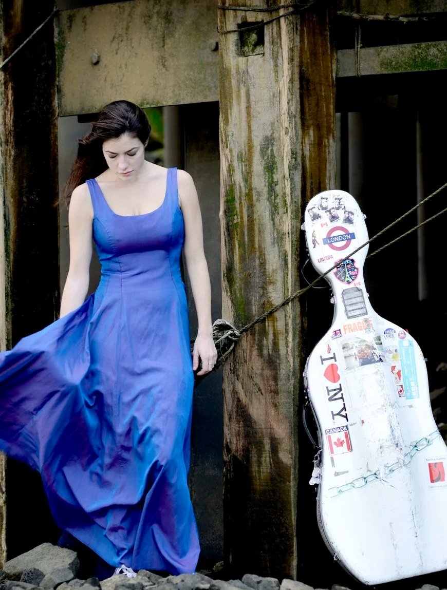 A woman in a blue dress standing outdoors near weathered wooden posts and a guitar case covered in stickers, including an 'I love NY' sticker.