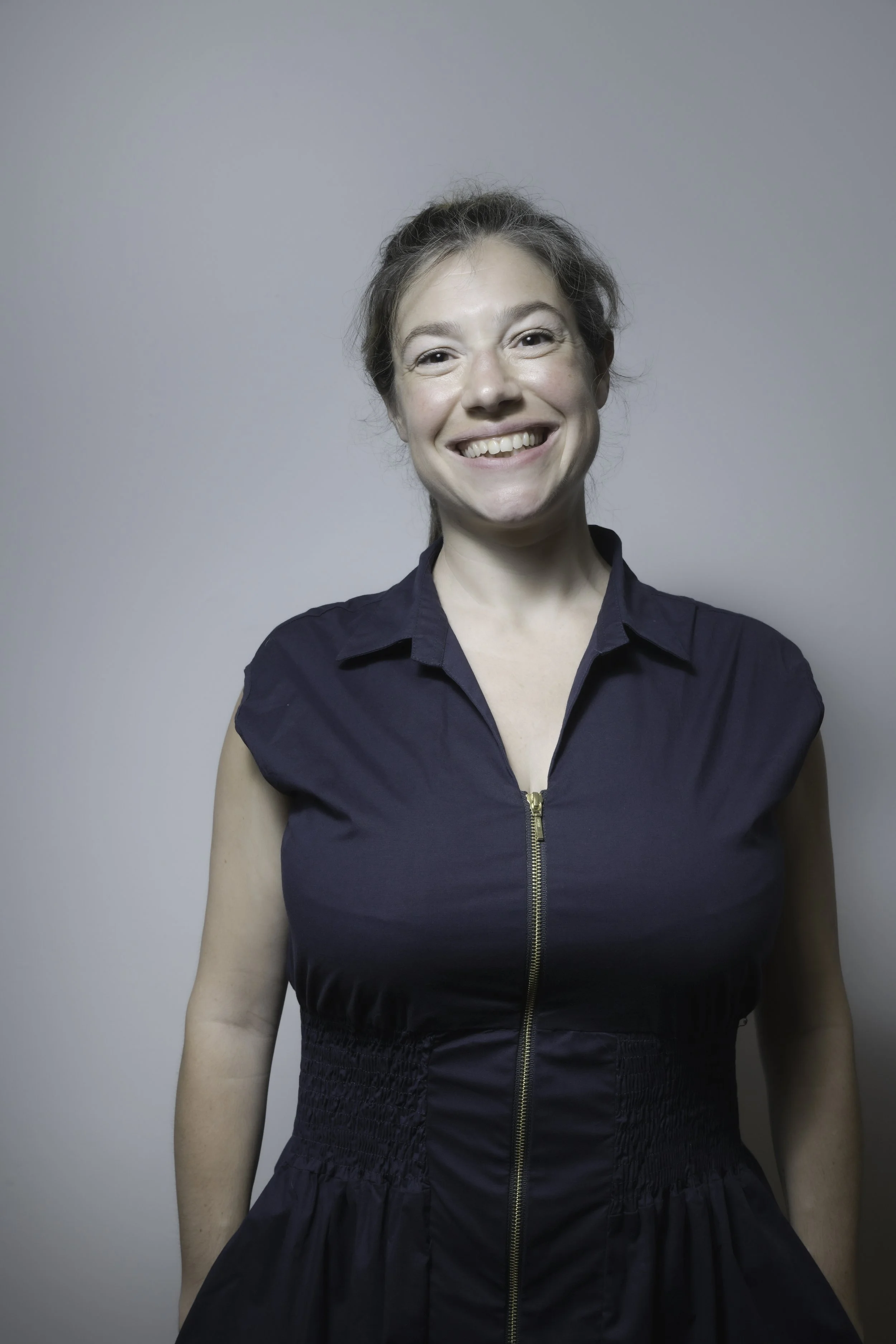 A woman with grayish hair smiling, wearing a sleeveless navy blue garment with a front zipper, standing against a plain light gray background.