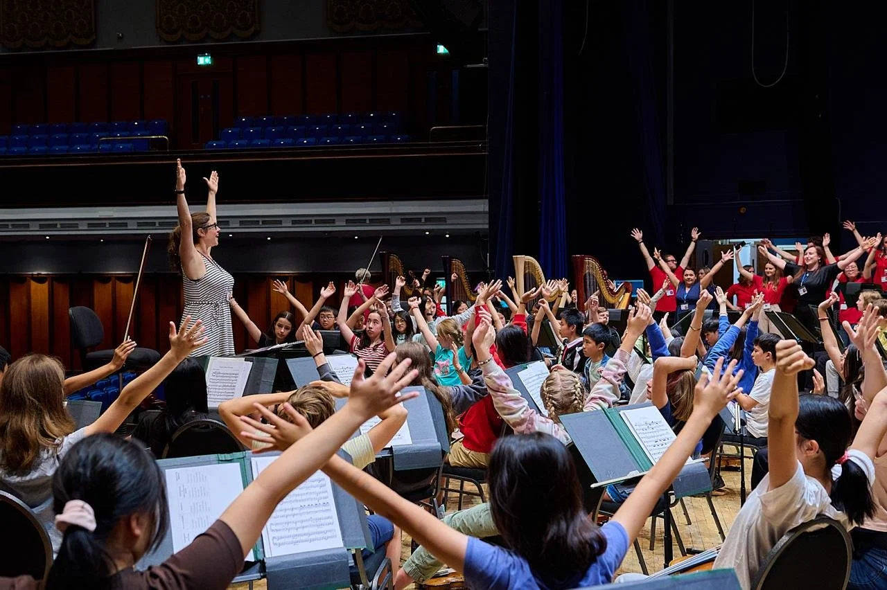 Orchestra rehearsal with children and a conductor on stage, raising their hands.
