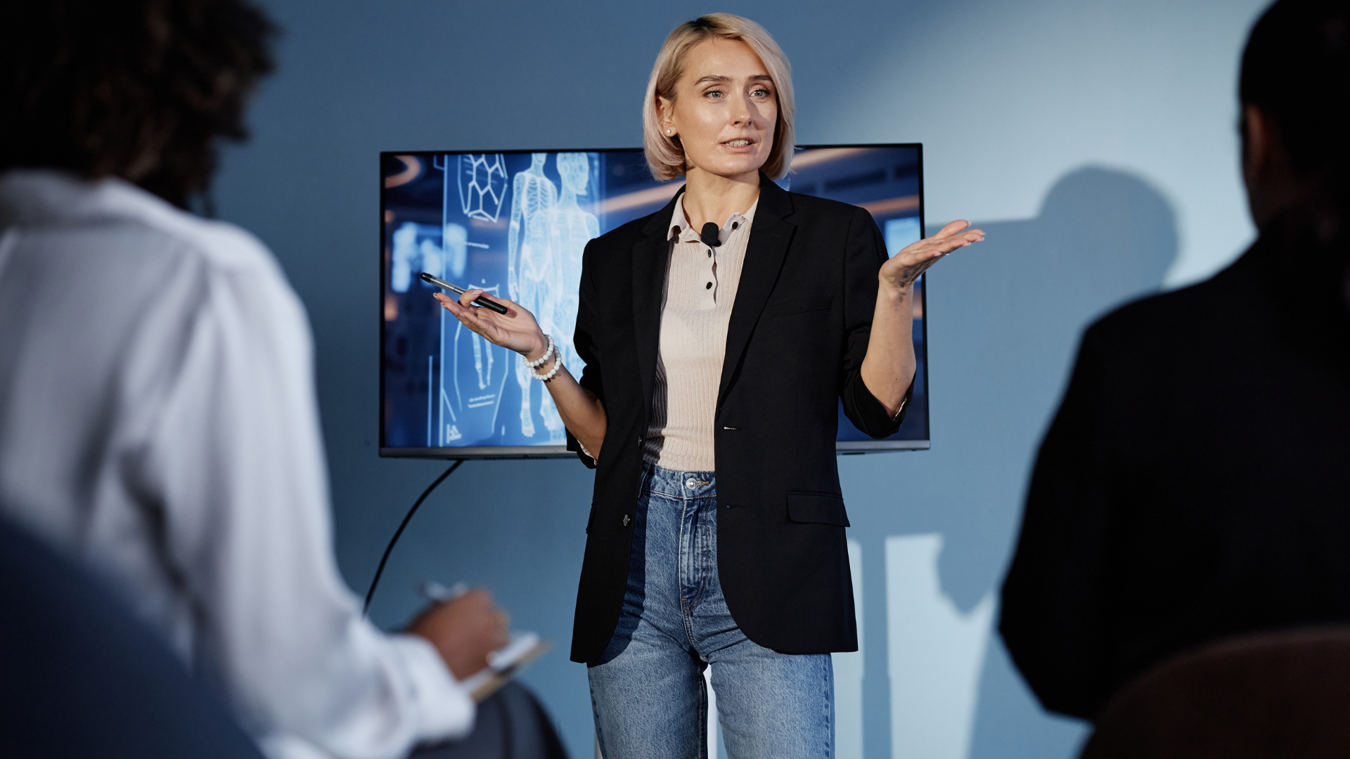 A woman in a black blazer and jeans giving a presentation about medical imaging to an audience in a conference room.