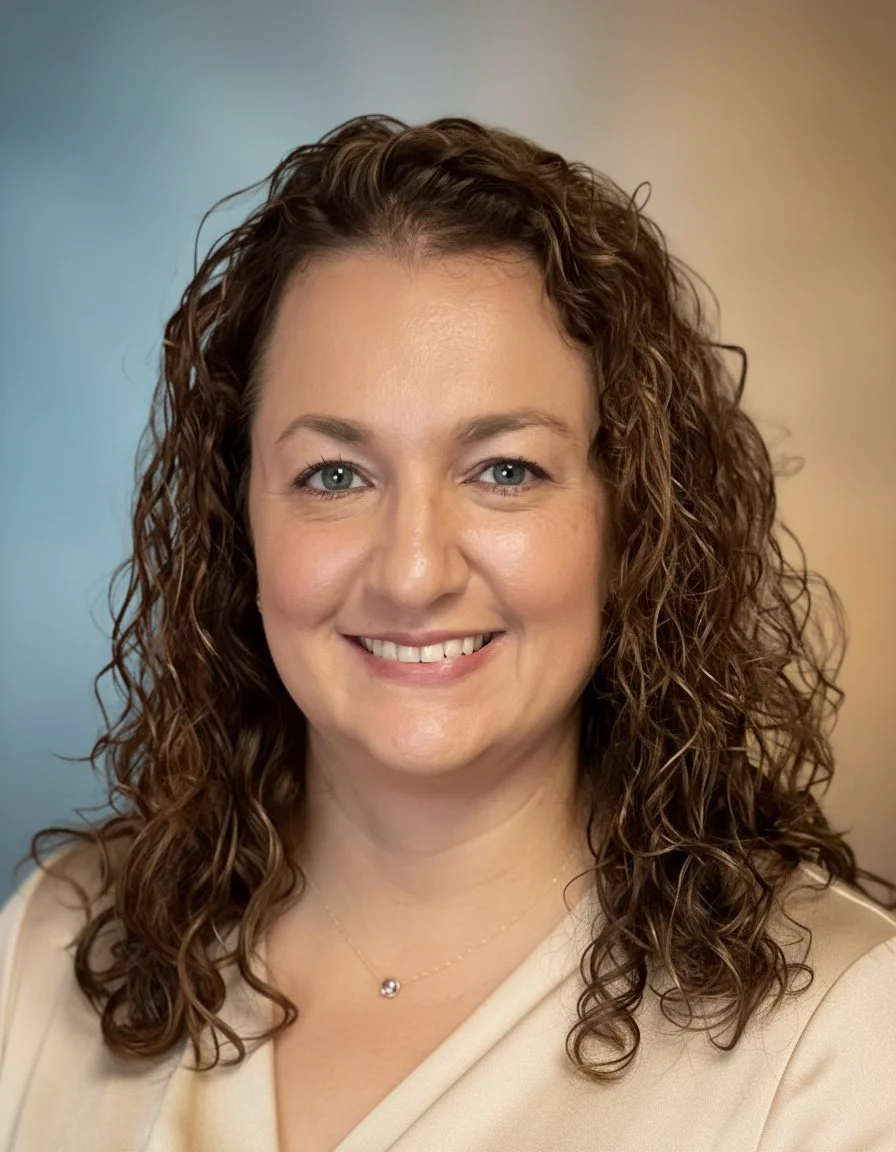 A smiling woman with curly brown hair and blue eyes, wearing a light-colored blouse and a delicate necklace, standing against a plain background.