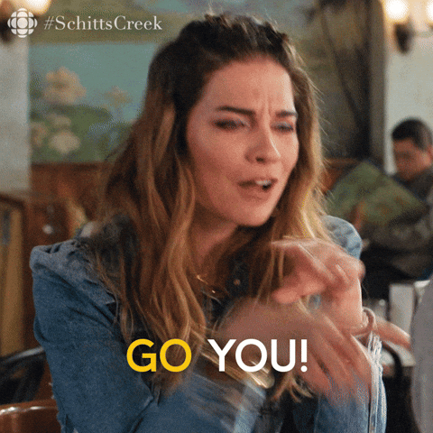 A woman with long brown hair making an animated expression, pointing towards the camera, in a restaurant setting. Text overlay says "GO YOU!" and a hashtag "#SchittsCreek" is visible in the top left corner.