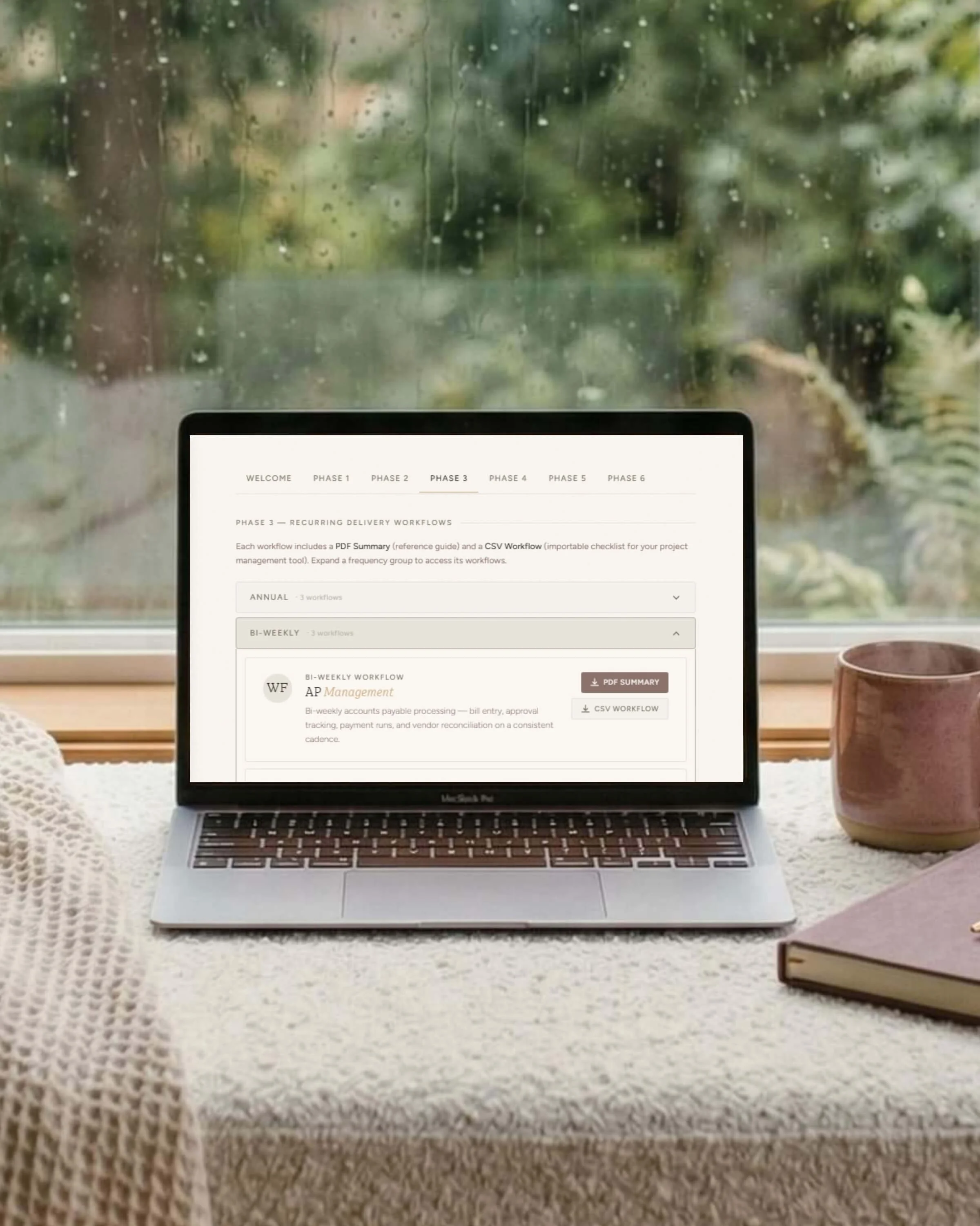 Open MacBook laptop on a table in front of a rain-streaked window, displaying a project management software. To the right of the laptop, there is a purple ceramic mug and a closed book with a purple cover. To the left, a white textured cloth and a small white decorative item are visible.