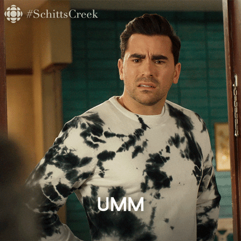 A man with dark hair and a serious expression looking to the side while wearing a white and black tie-dye sweater inside a room with wooden and teal elements.