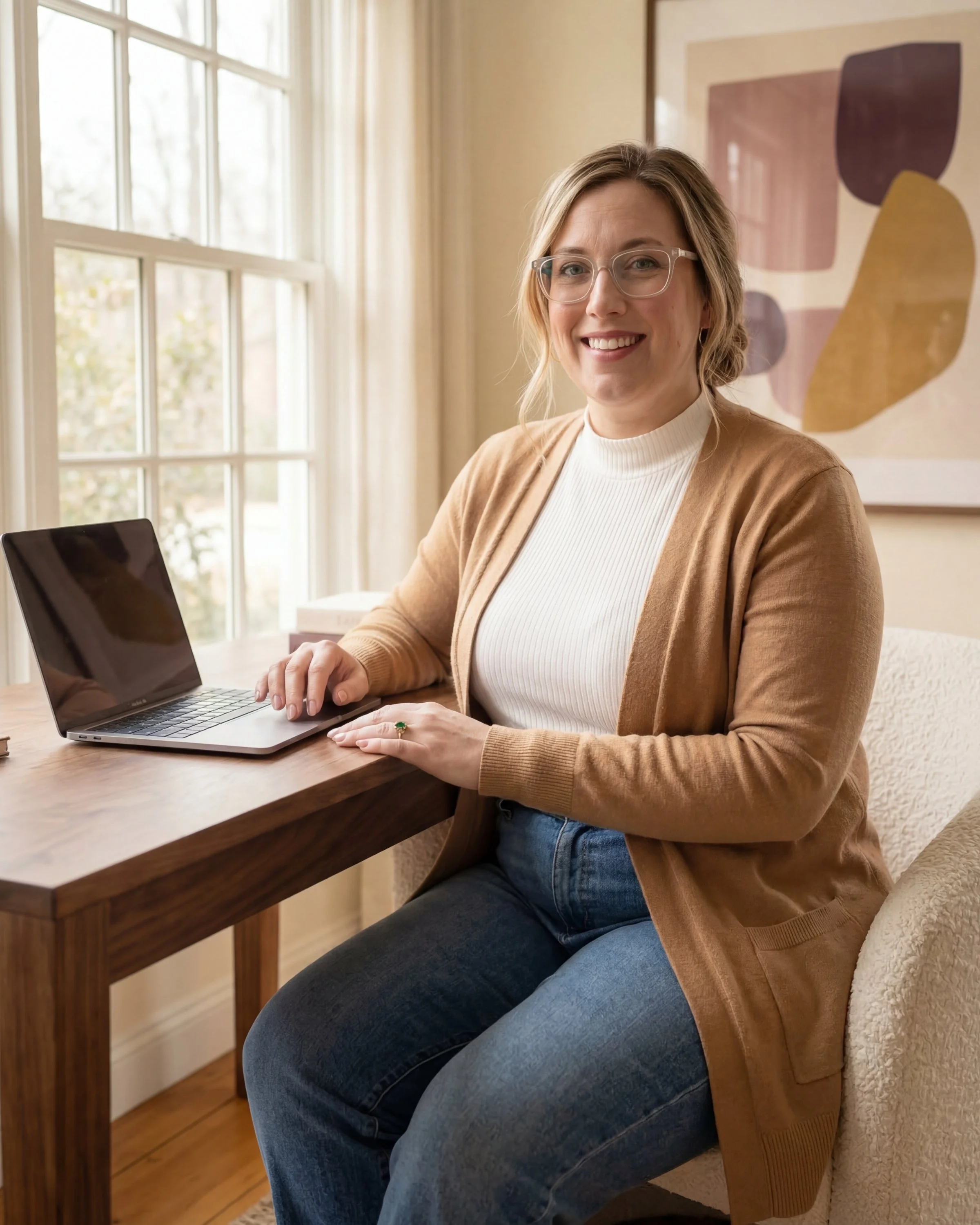 A woman with glasses sitting at a wooden desk with a laptop, smiling at the camera.