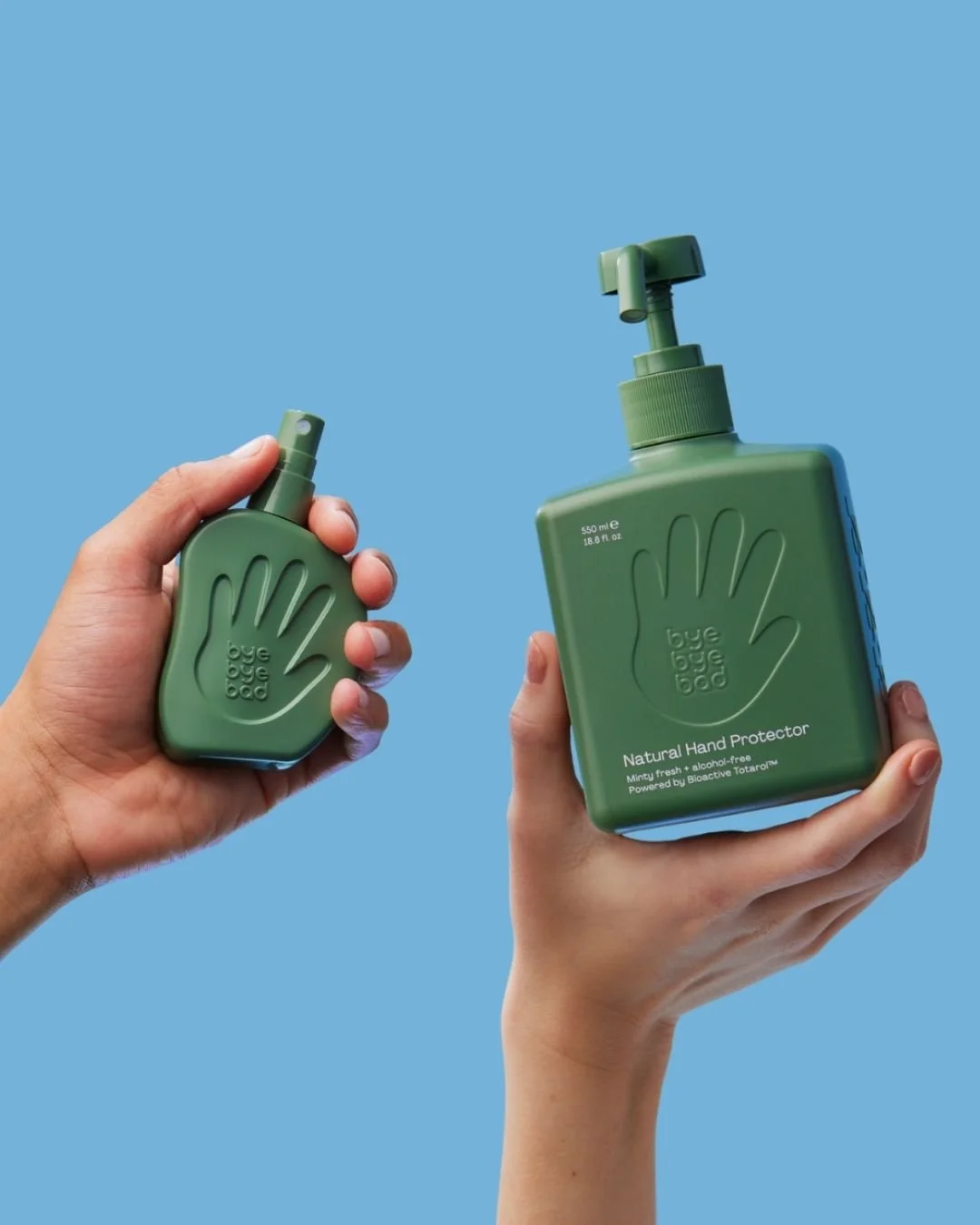 Two hands hold green bottles of hand sanitizer against a blue background, one spray bottle and one pump bottle, both labeled 'bye bye bad' and 'Natural Hand Protector'.