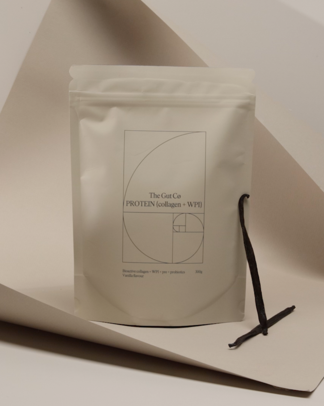 A white resealable bag labeled "The Gut Co Protein (collagen + WPI)" with a minimalist geometric design, placed on white paper with a dark vanilla bean pod beside it.
