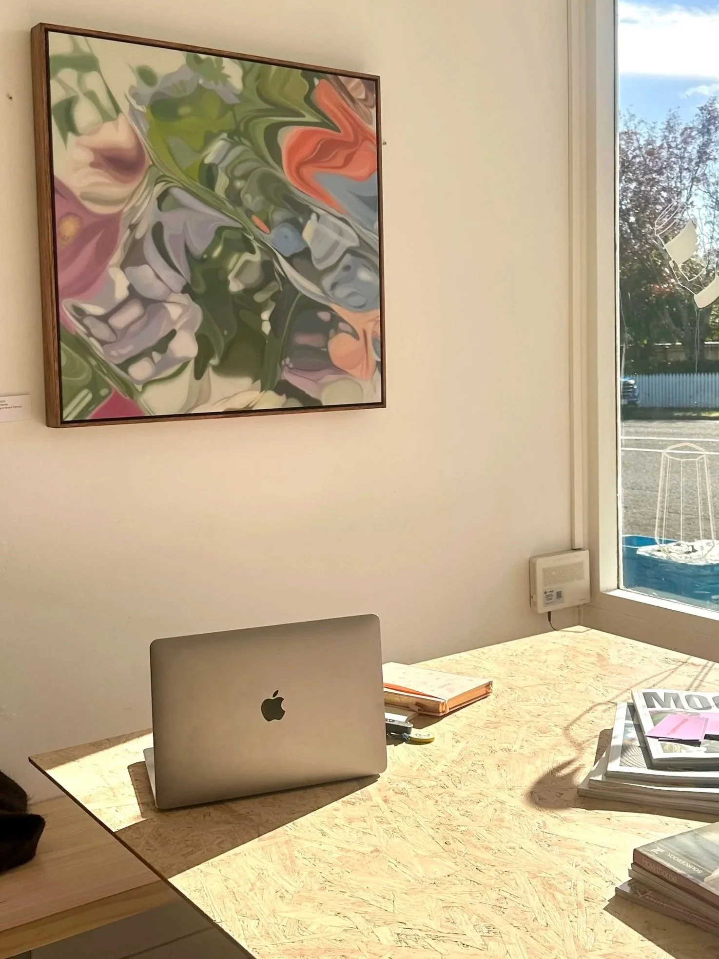 A wooden desk with an Apple MacBook, a stack of magazines, notebooks, and pens inside a well-lit room with a large abstract painting on the wall and a window showing trees outside.