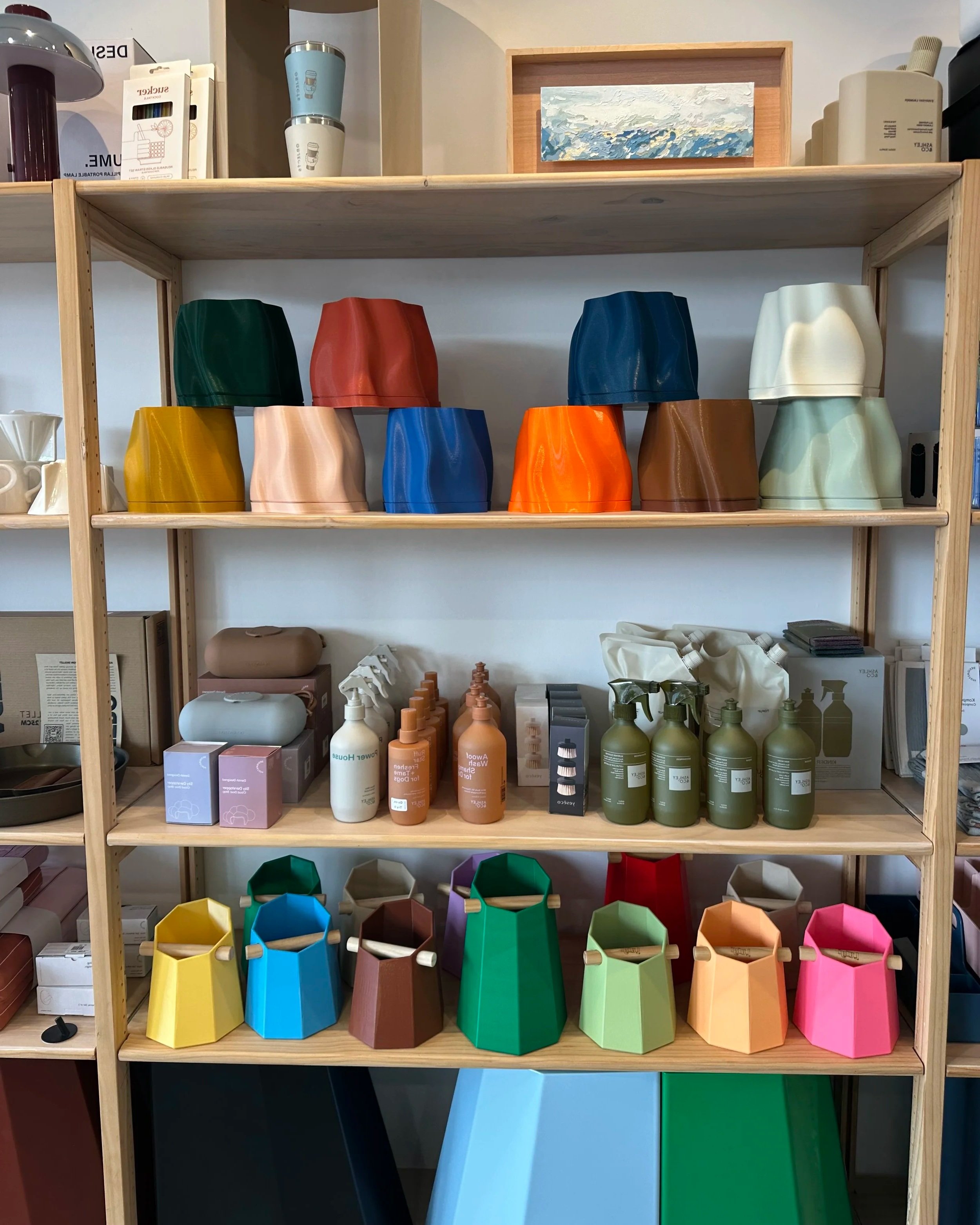 Colorful ceramic or plastic vases and containers on wooden shelves, with the top shelf holding cups and small containers, and the bottom shelf displaying aromatic bottles and dispensers, possibly in a home decor or gift shop.