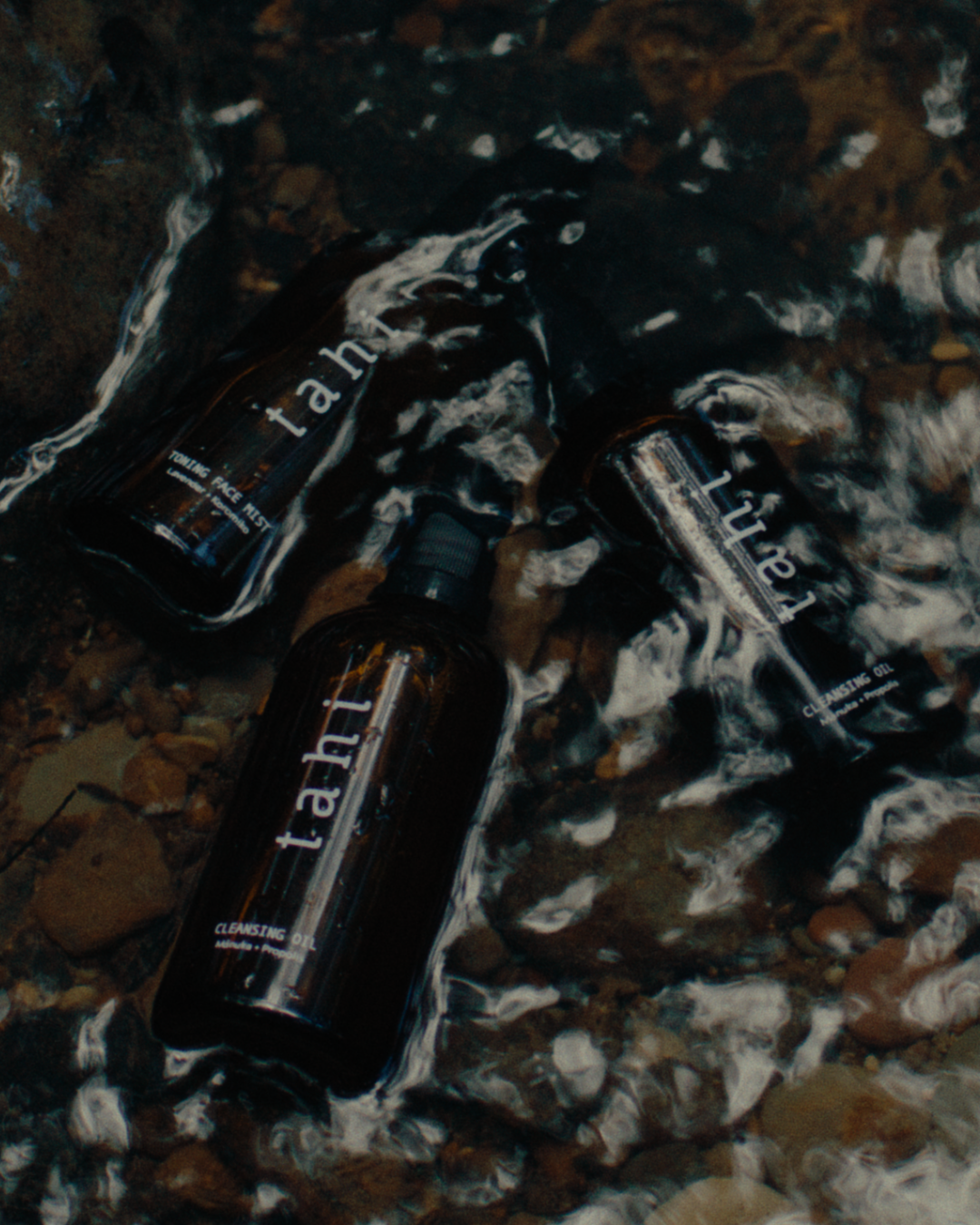 Three bottles of skincare products, including toner and cleansing oil, submerged in water on a rocky surface.