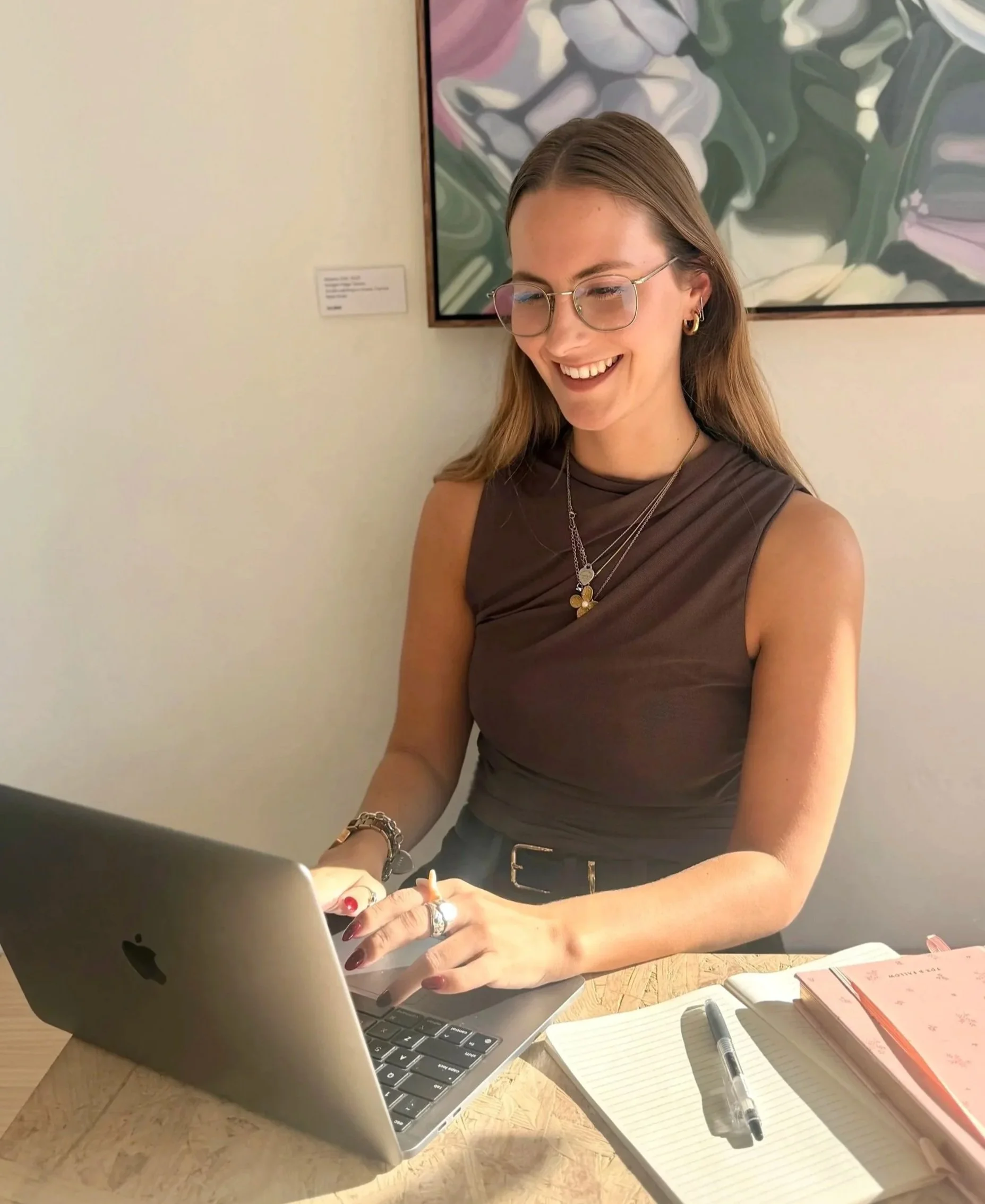 A woman with long light brown hair, glasses, and gold jewelry is smiling while working on a silver MacBook laptop at a desk. There are open notebooks and a pen in front of her. She is wearing a sleeveless brown top and has a large painting hanging on the wall behind her.