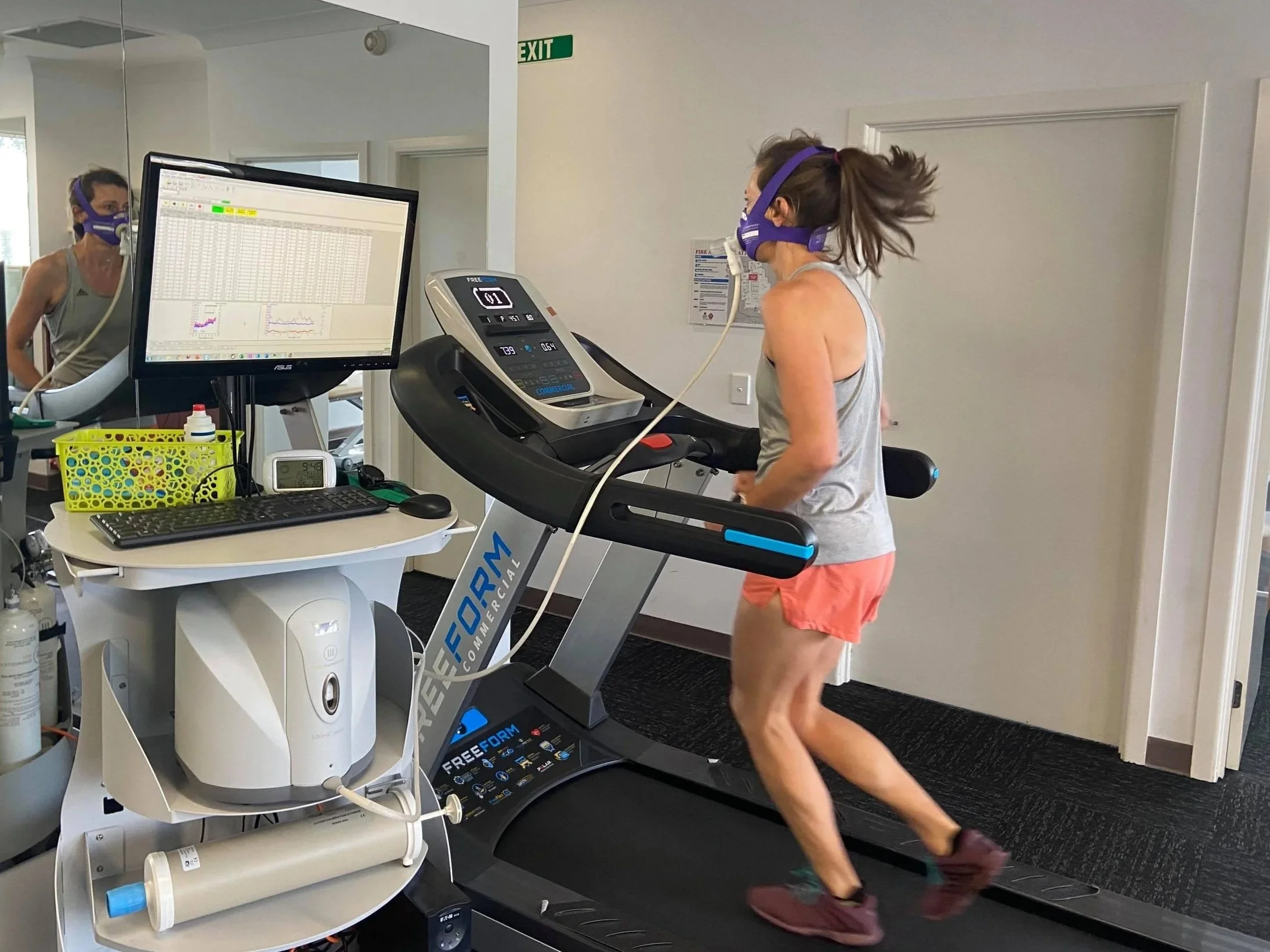A woman running on a treadmill in a gym, wearing a purple mask, gray tank top, and orange shorts. She is focused on a monitor displaying heart rate and other data. There is a mirror and a computer with a large screen nearby, showing graphs and data.