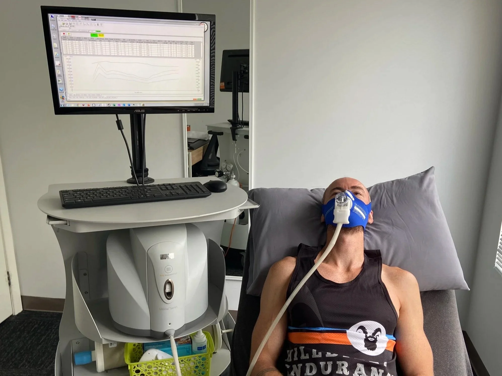 A man lying on a bed or examination table with oxygen mask on his face, connected to medical equipment, in a clinical setting.