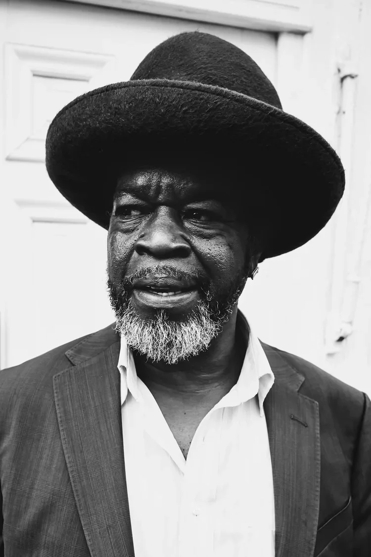 Black and white photo of a man with a beard wearing a wide-brimmed hat and a suit jacket, standing outdoors in front of a building.