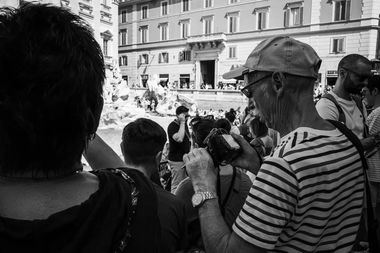 A crowded outdoor scene with people gathered, some looking at a man holding a camera, in front of a historic building with ornate architecture.