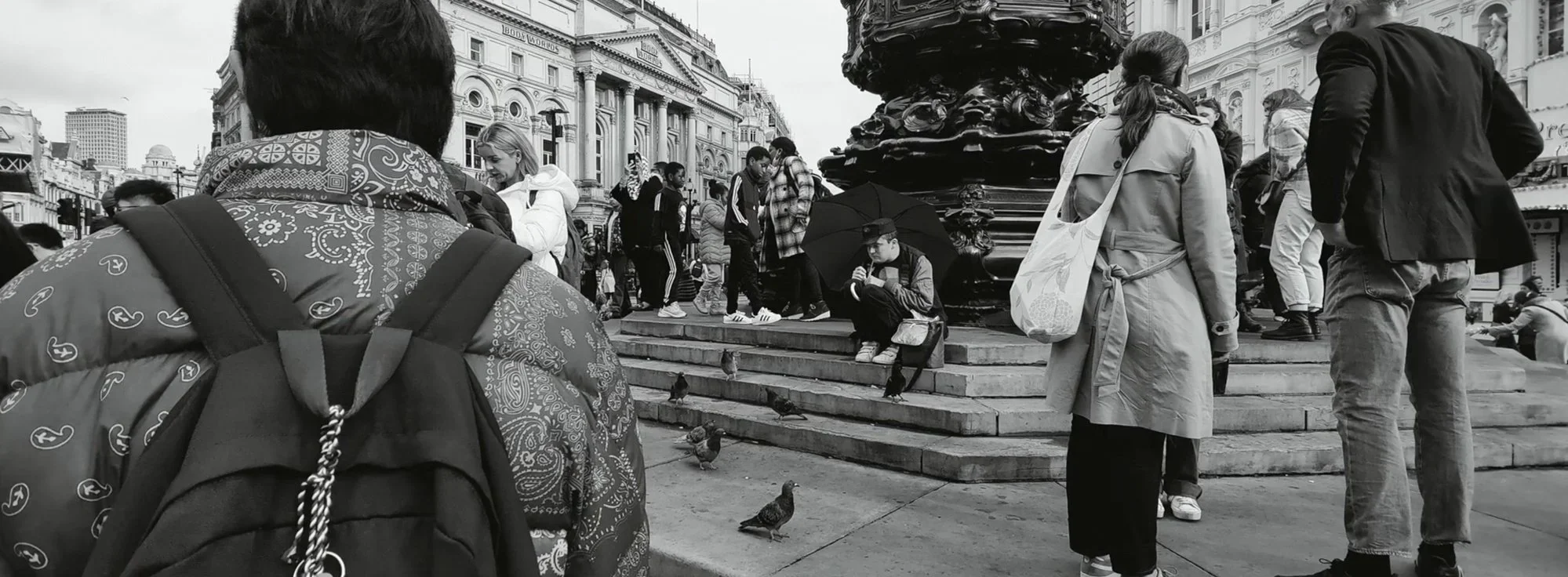 Crowded city square with people walking, sitting, and pigeons on the steps near a large ornate fountain, with historic buildings in the background, black and white photo.