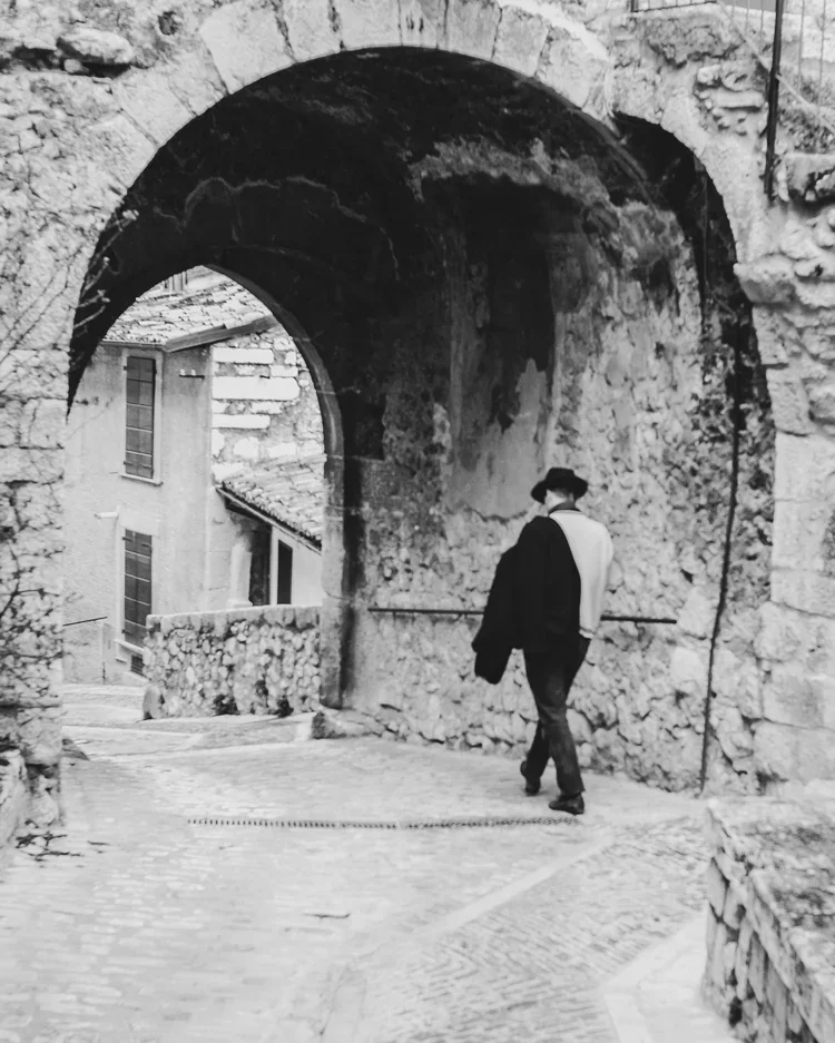 A person dressed in dark clothing and a hat walking through an old stone archway in a historic area.