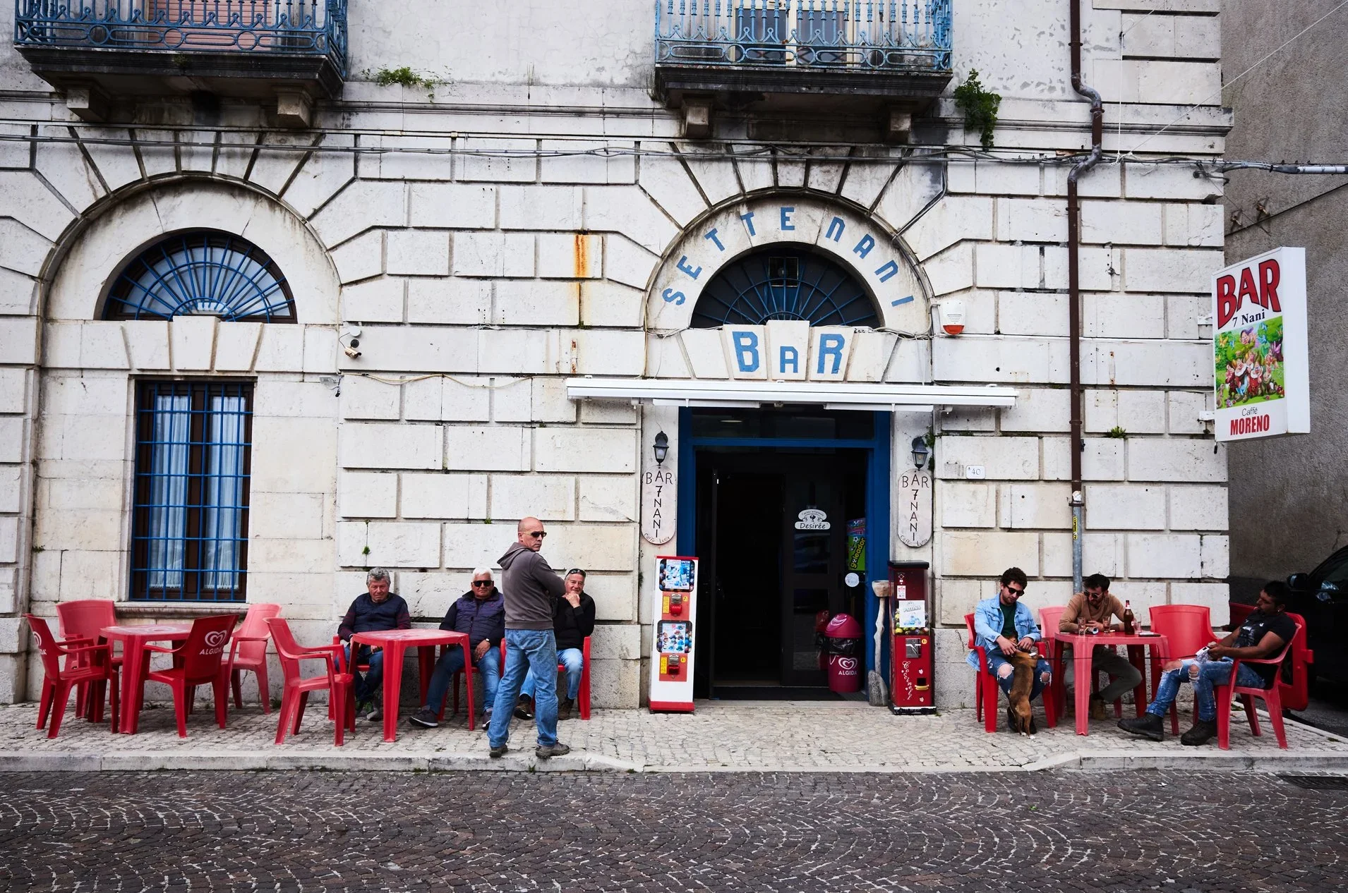 People sitting outside a bar with red tables and chairs on a cobblestone sidewalk, with a white stone building with arched windows and a blue sign above the entrance that reads 'SETE NANI' and a smaller sign that says 'BAR', a vending machine, and a 