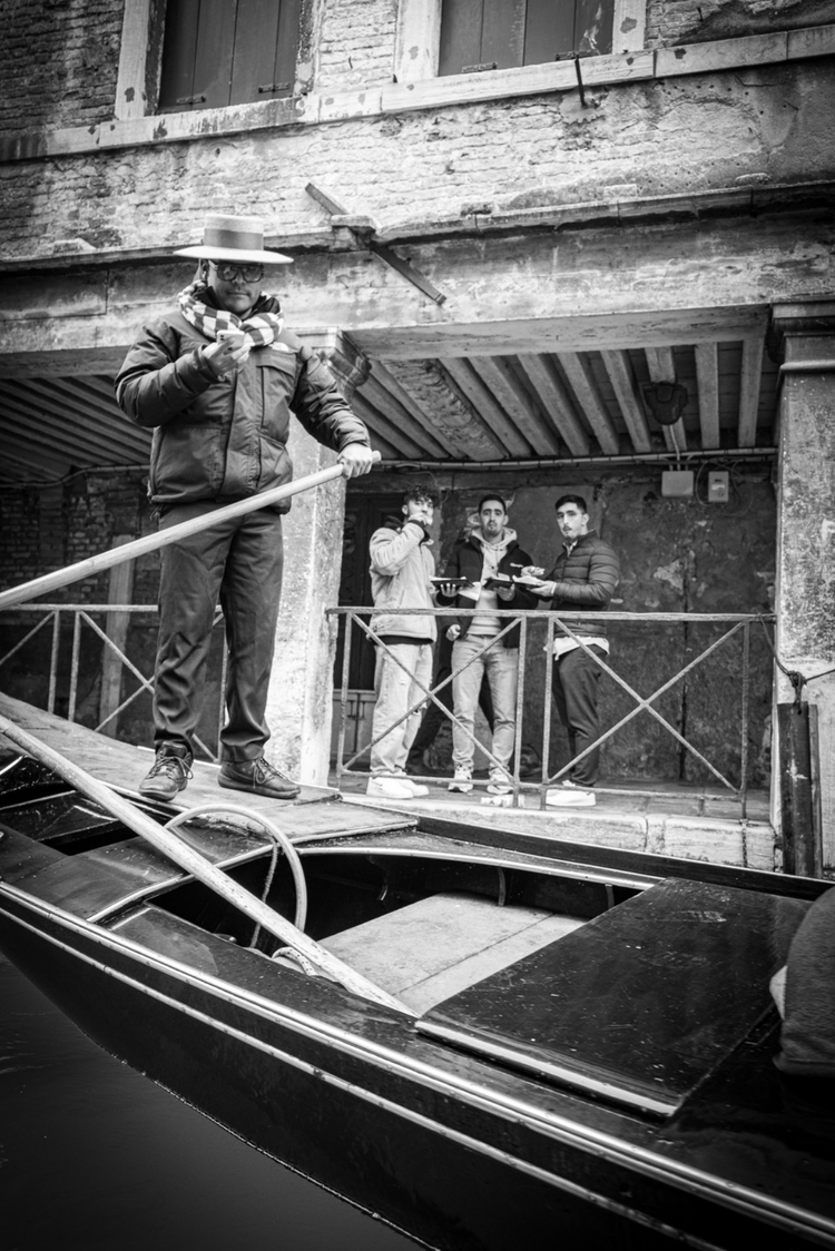 Four men on a dock next to a boat, with three standing on the dock holding plates of food. One man wearing a hat and scarf is on the boat holding a pole and looking at his phone.