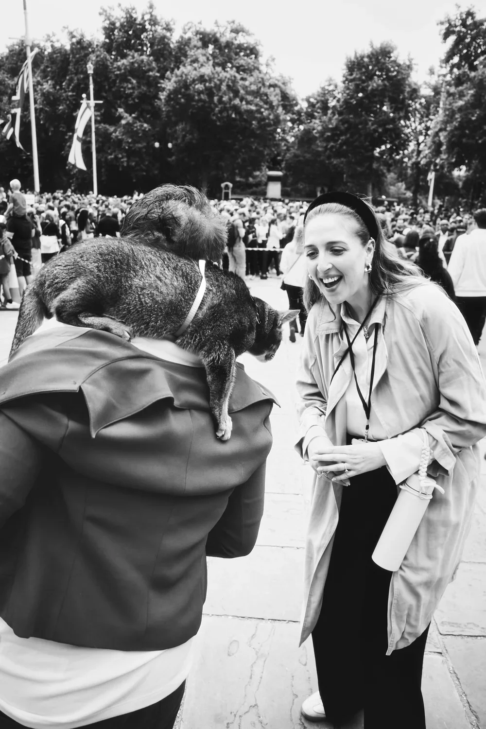 A woman laughing and talking to a person with a dog on their shoulders at an outdoor event with a large crowd in the background.