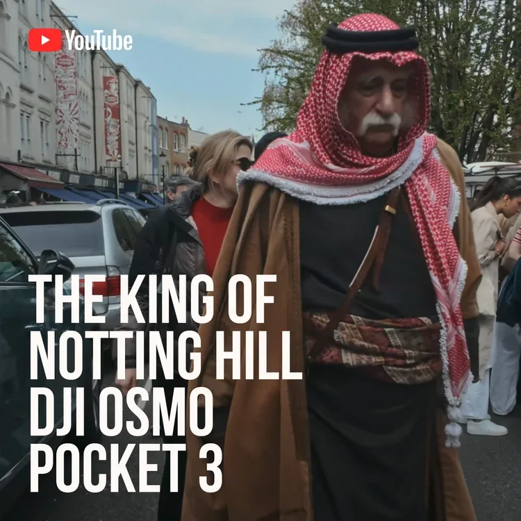 A man dressed in Middle Eastern attire, wearing a red and white checkered keffiyeh and a brown robe, walking outdoors among a crowd of people on a city street with cars and buildings in the background.