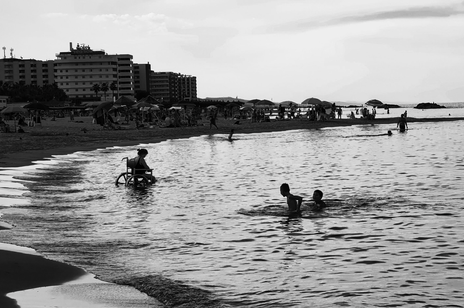 People enjoying a beach with umbrellas, some in the water, and buildings in the background, black and white photo.