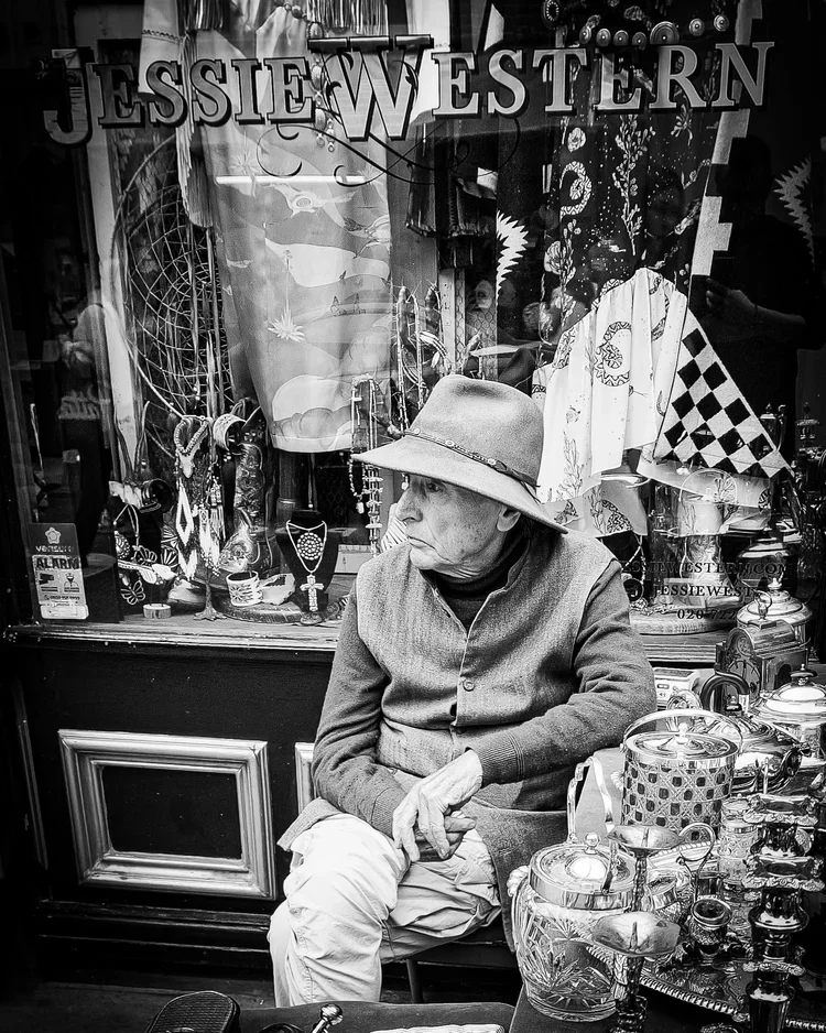 An elderly man wearing a hat and a jacket sits outside a shop window displaying jewelry and textiles. The scene is in black and white.