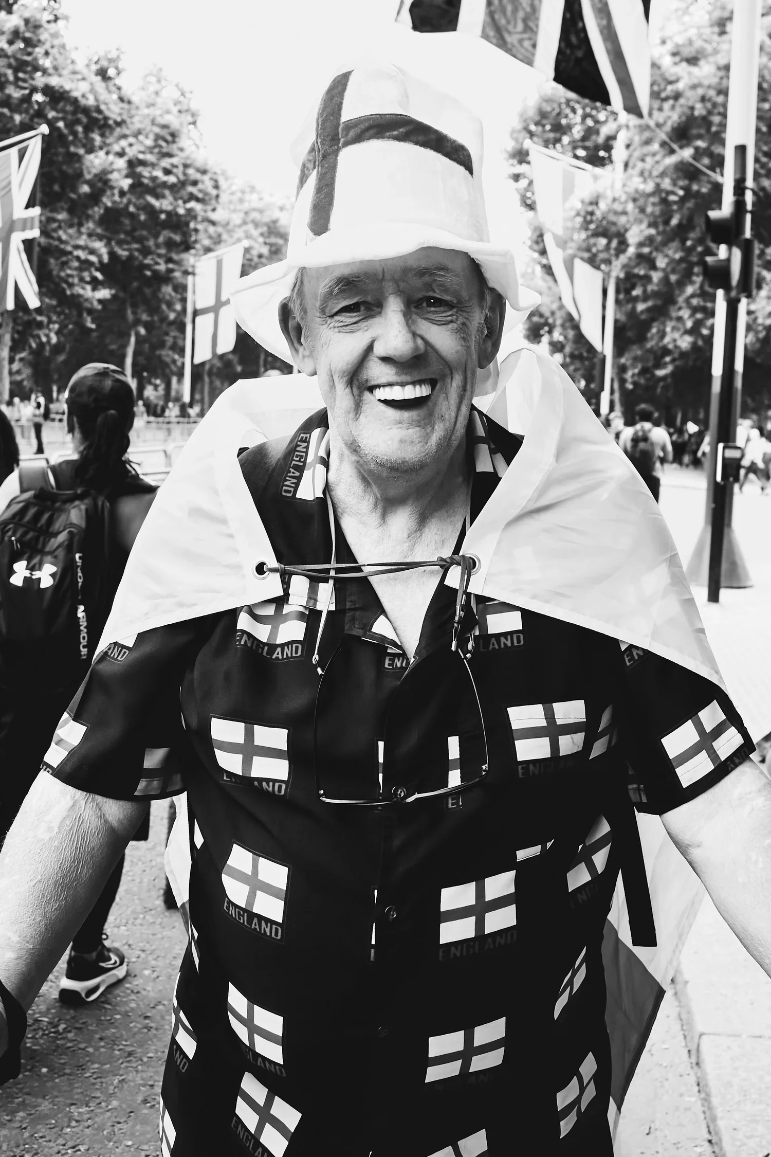 A smiling man wearing an England-themed shirt and a large England hat, with an England flag draped over his shoulders, standing outdoors among other people and flags.