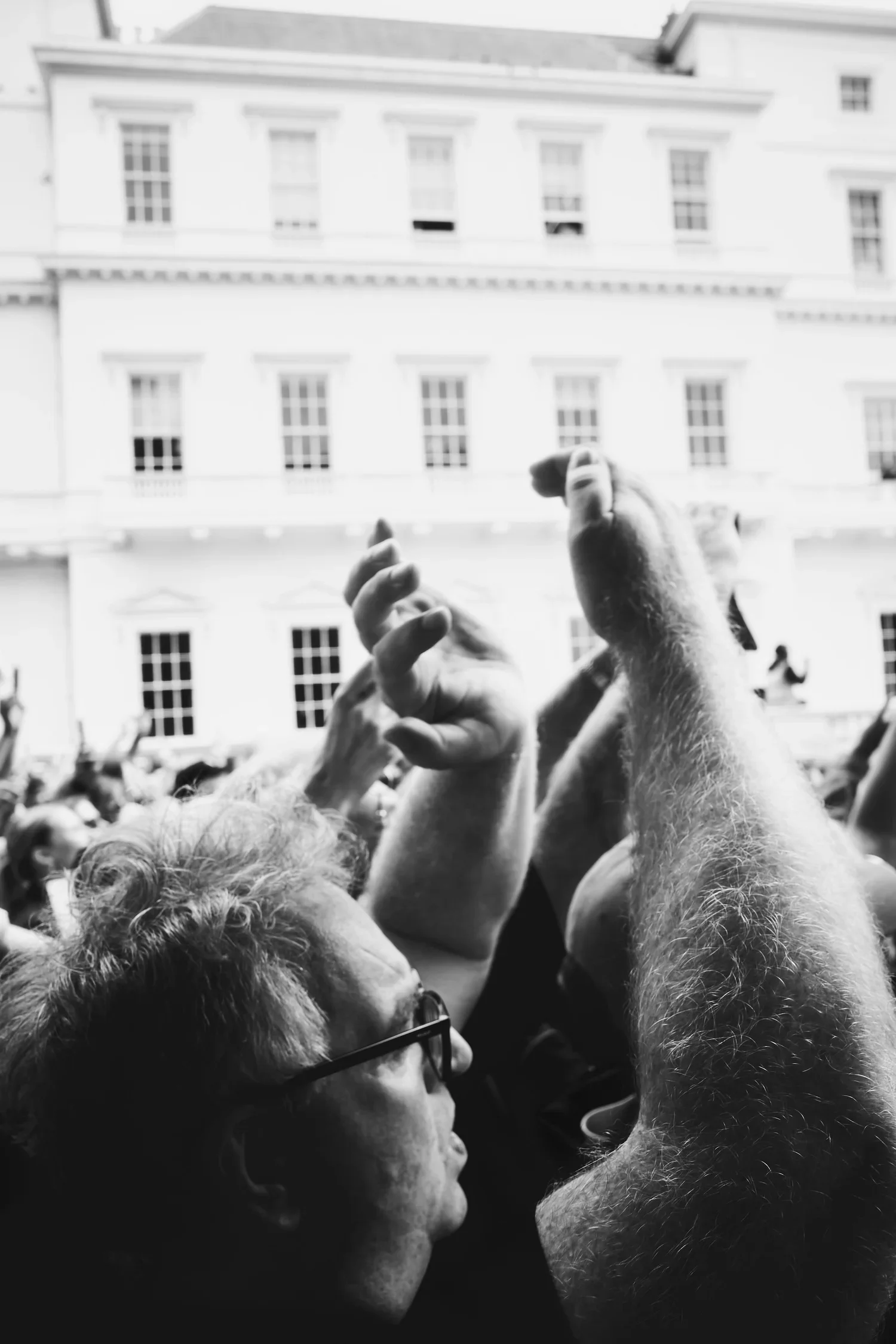 Crowd of people at a concert, with a woman with glasses and curly hair in the foreground, and individuals raising their hands around her, with a large building with multiple windows in the background.