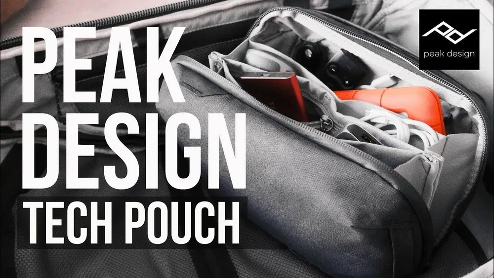A gray tech pouch inside a backpack containing various electronic accessories and gadgets, with large white text overlay reading "Peak Design Tech Pouch" and a small Peak Design logo in the top right corner.