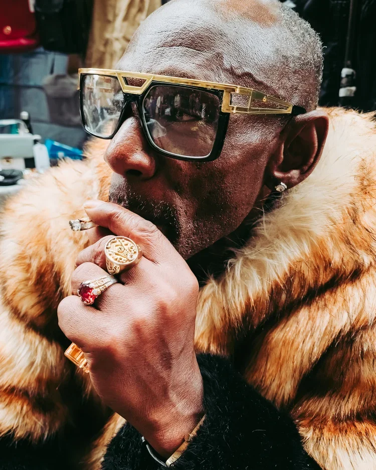 A man with gray hair, wearing stylish large sunglasses and a fur coat, is smoking a cigar with rings on his fingers.
