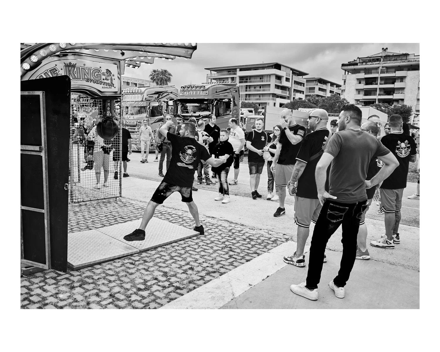A group of men and boys gathered outdoors around a carnival game where a young man is throwing a ball at a mounted object. Several trucks and mid-rise buildings are in the background.