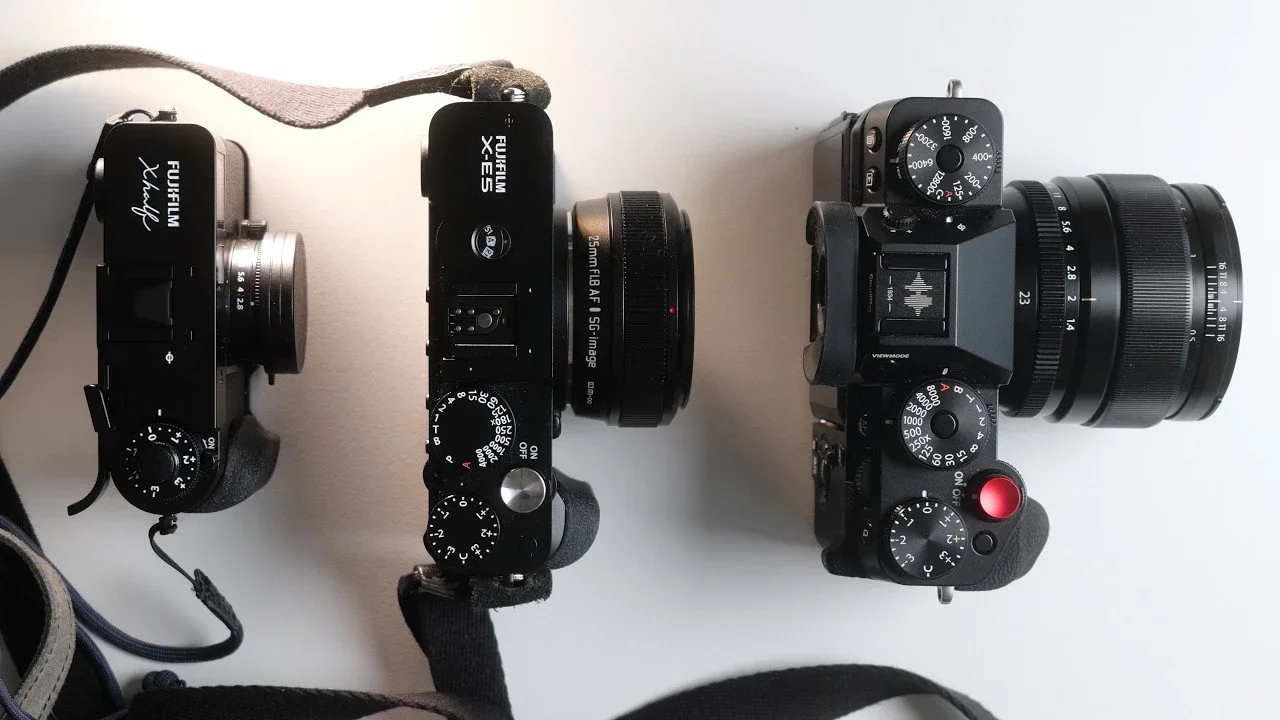 Three digital cameras with black bodies and attached lenses are arranged on a white surface.