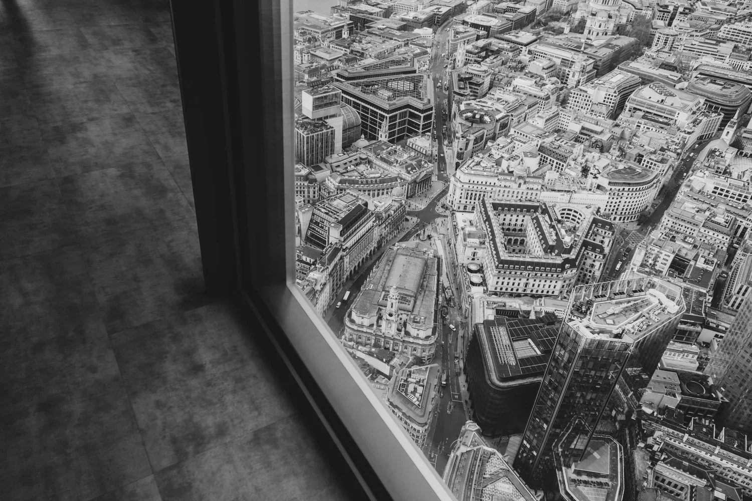 A black and white photo taken from inside a high-rise building, looking out through a window at a dense cityscape with numerous tall buildings, streets, and rooftops.
