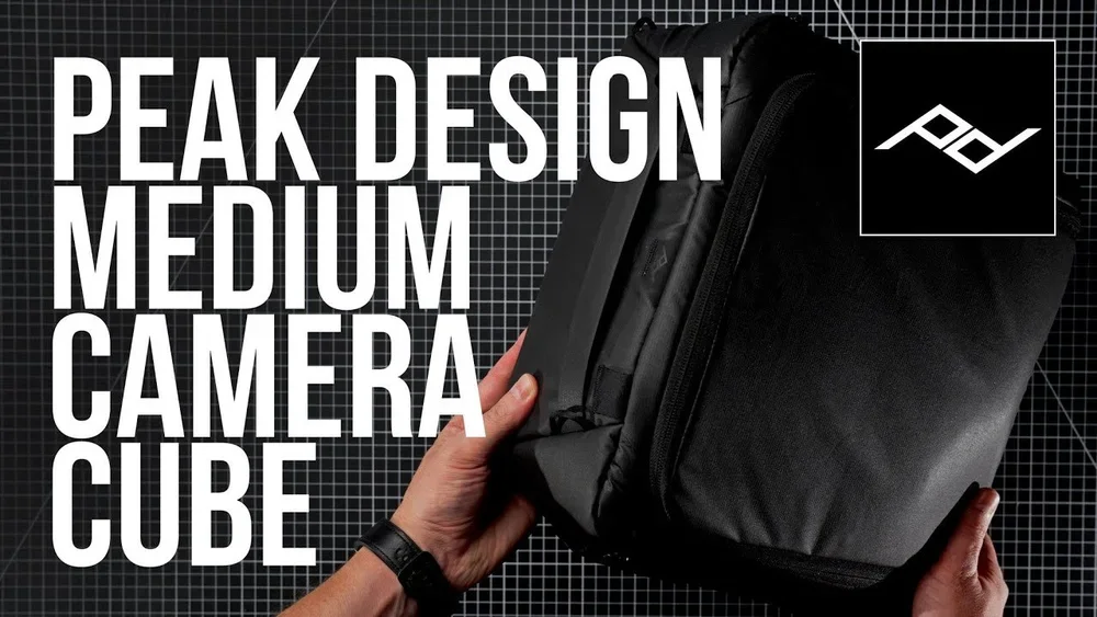 A person holding a black backpack with large white text on a black background that reads "PEAK DESIGN MEDIUM CAMERA CUBE".