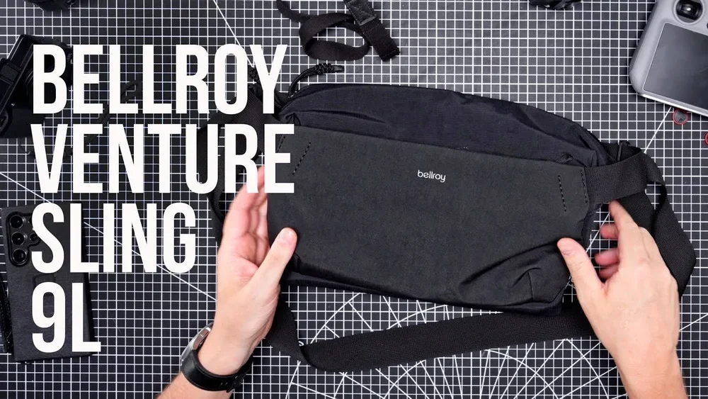 Hand holding a black Bellroy Venture Sling 9L bag on a black and white grid surface surrounded by a camera and accessories.