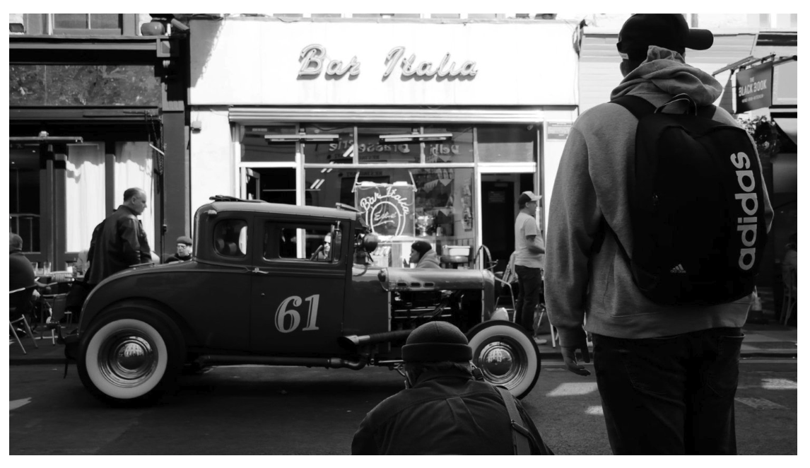People gathered on a city street near a vintage race car with the number 61, in front of a cafe with a sign that reads 'Bar Italia'.
