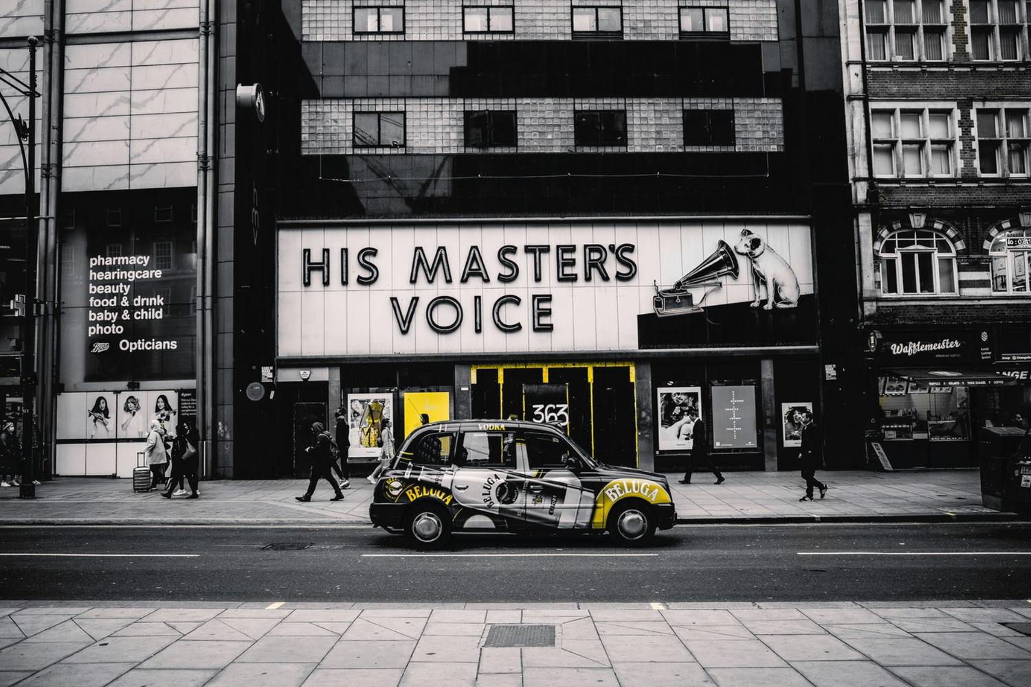 Black and white city street scene featuring a vintage black taxi with yellow accents parked on the road. In the background, a large illuminated sign reads 'HIS MASTER'S VOICE' with a picture of a dog listening to a gramophone. Pedestrians walk along 