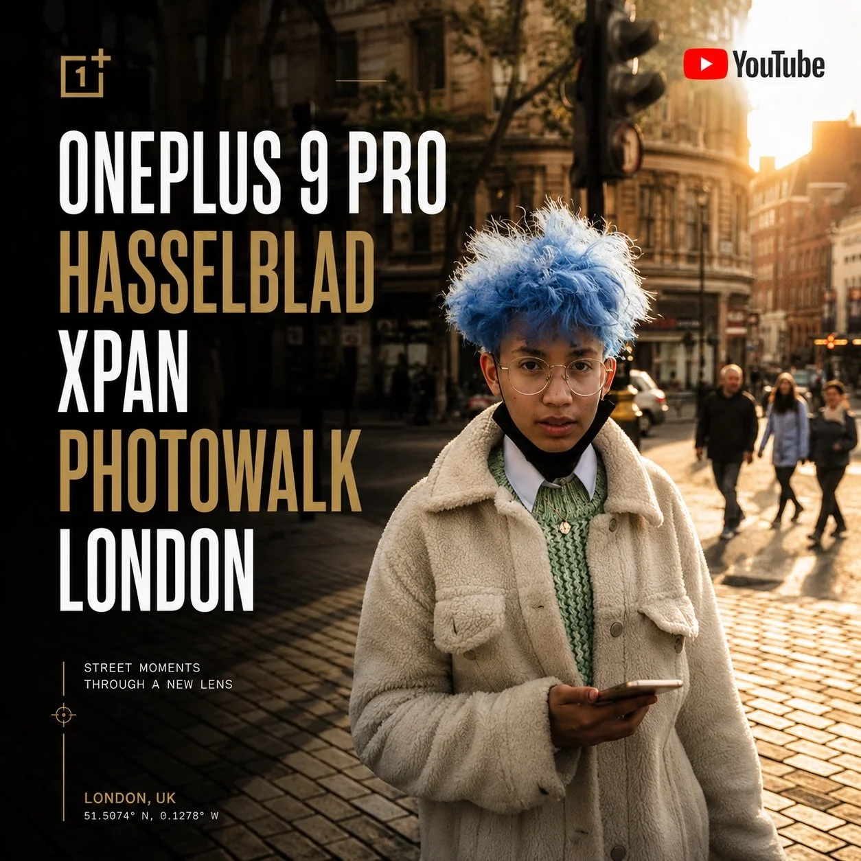 A person with blue hair, glasses, wearing a cream-colored jacket and green sweater, stands on a city street holding a phone, with a background of buildings, crosswalk signal, and pedestrians.