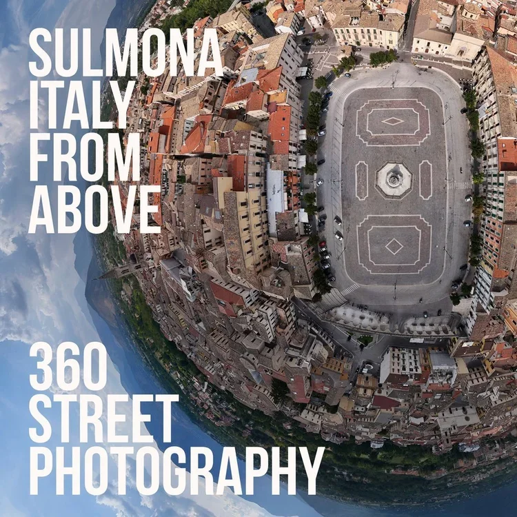 Aerial view of a town in Sulmona, Italy, showing a central square with a fountain, surrounded by tightly packed buildings and an open area with sports lines, under a cloudy sky.