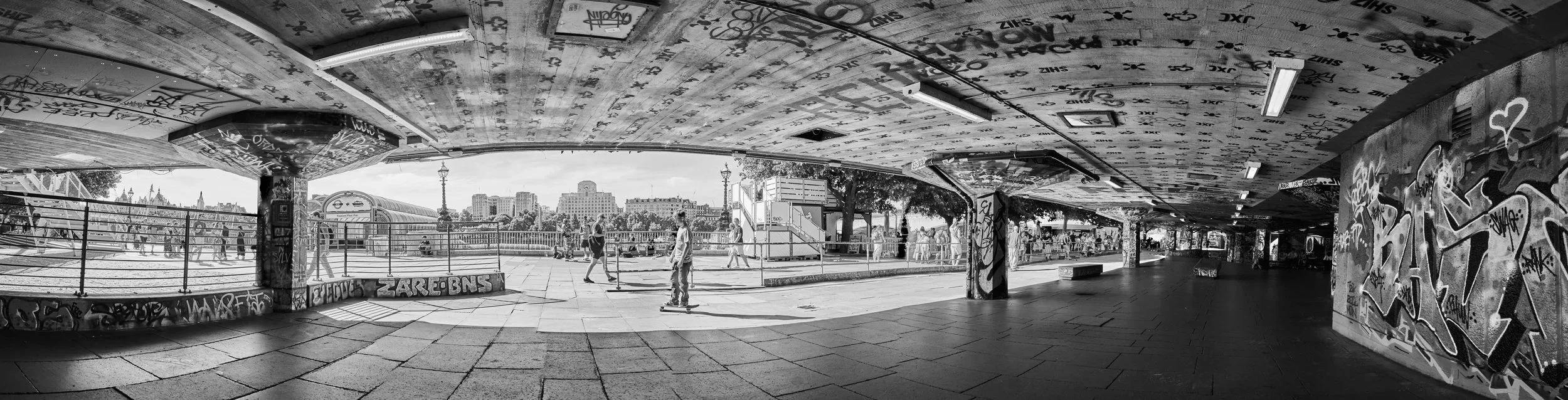 Underpass with graffiti-covered walls, graffiti on ceiling, and people skateboarding and walking outside on a sunny day.