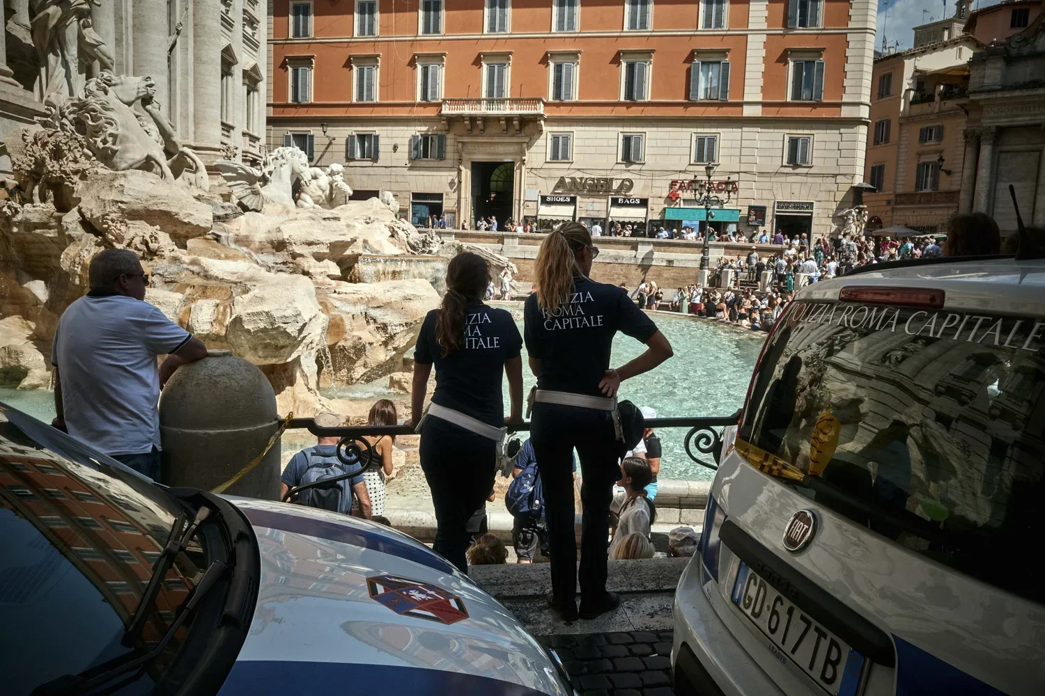 Tourists and police officers at the Trevi Fountain in Rome, Italy, with a large crowd and historic buildings in the background.