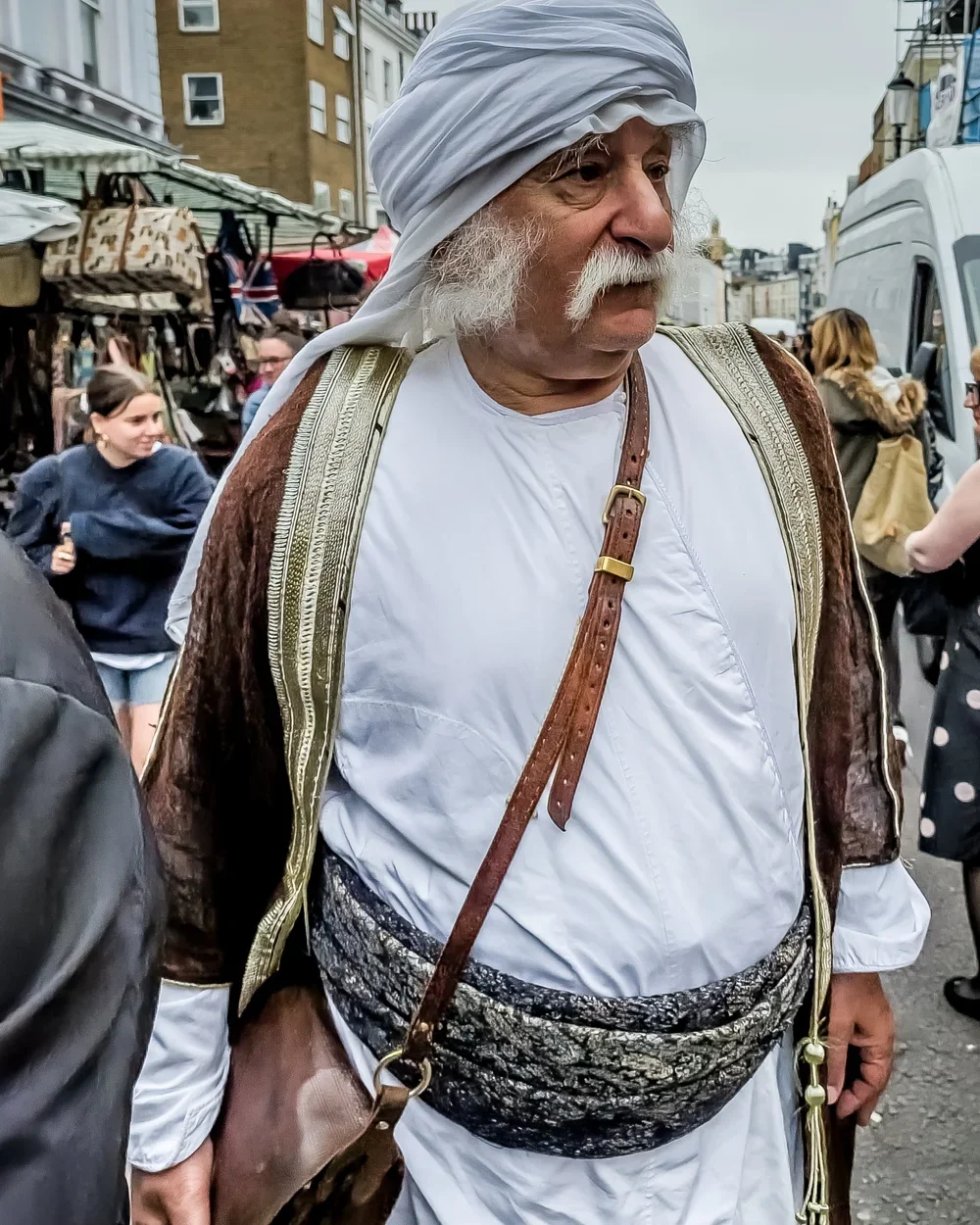 A man dressed in traditional Middle Eastern or North African attire, including a white turban and white garments, standing on a busy street market.