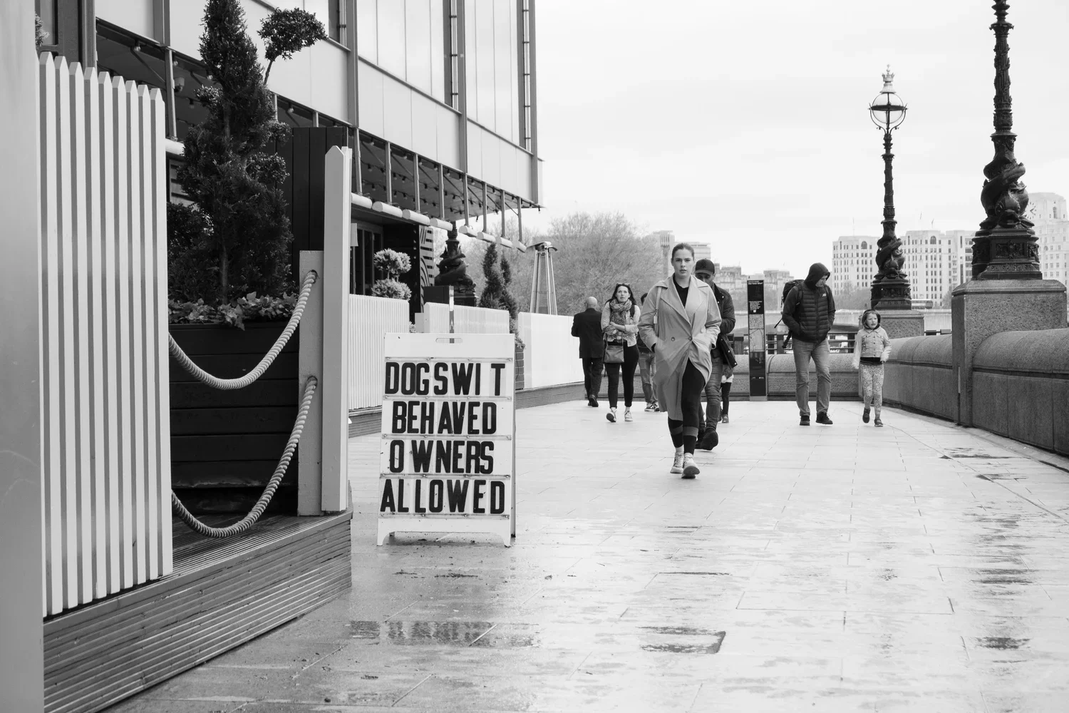 People walking on a city sidewalk near a sign that reads 'Dogs with Behaved Owners Allowed' in black and white, with a bridge railing and cityscape in the background.