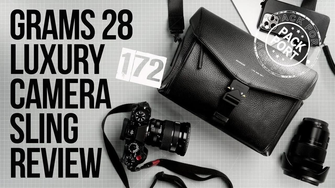 Black leather camera bag, camera with lens, additional camera lens, camera strap, and a sticker with 'pack port' logo, laid out on a grey grid surface.