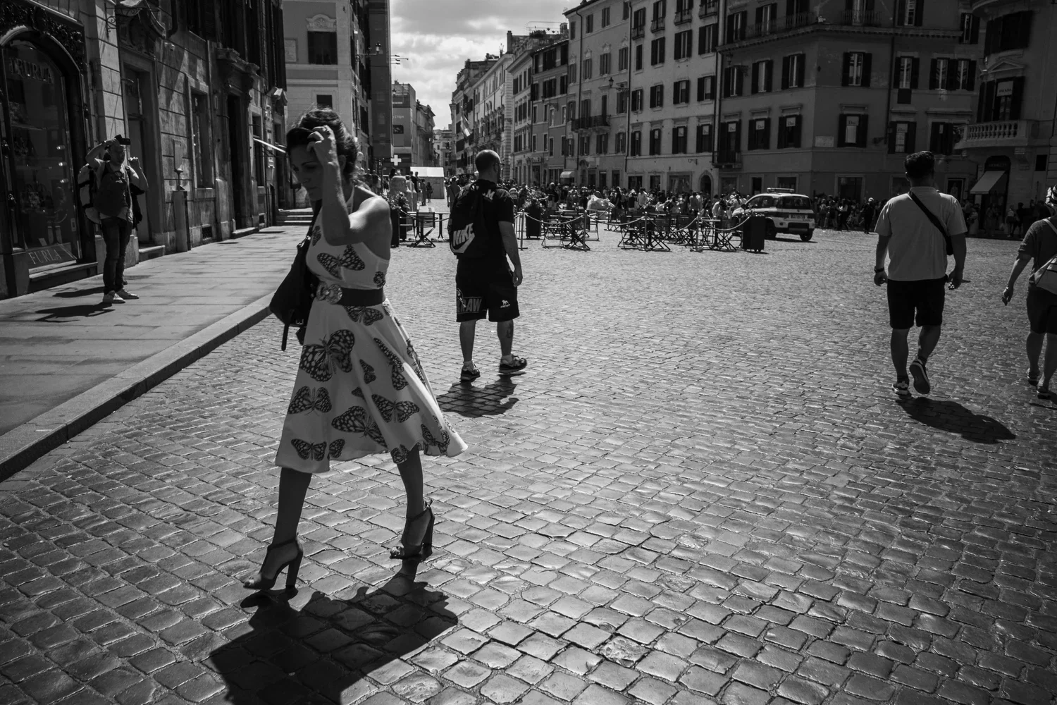 A black and white photo of a busy European city street with multiple pedestrians, including a woman in a butterfly-printed dress and high heels, a man in a shorts, and others walking or standing. There are old buildings with shopfronts lining the str