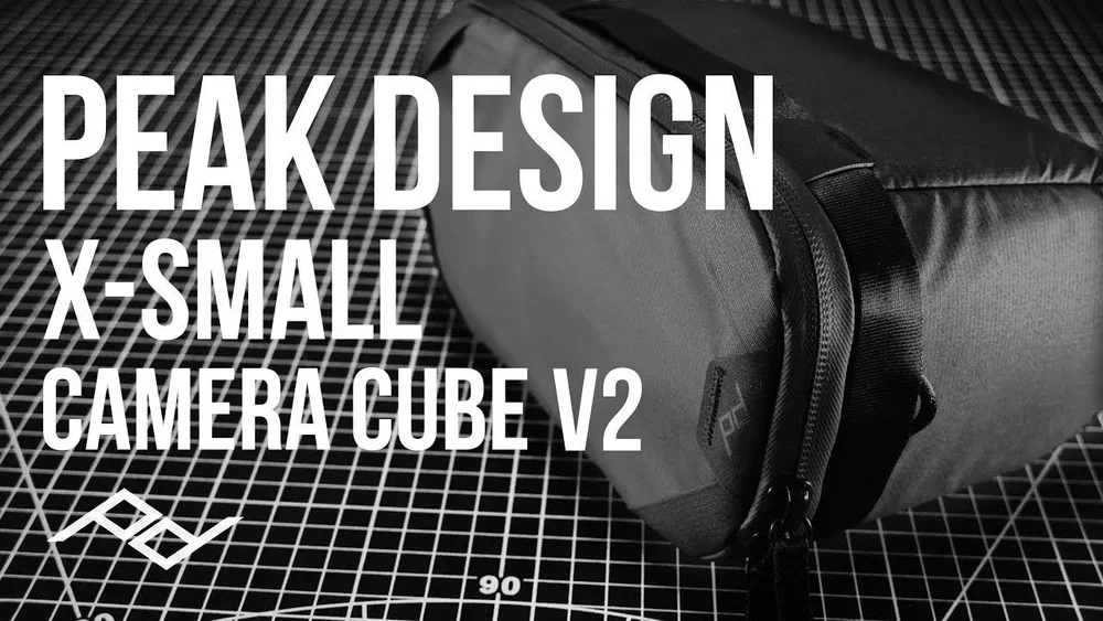 A black camera bag placed on a cutting mat with grid lines. Overlaid text announces the Peak Design X-Small Camera Cube V2.