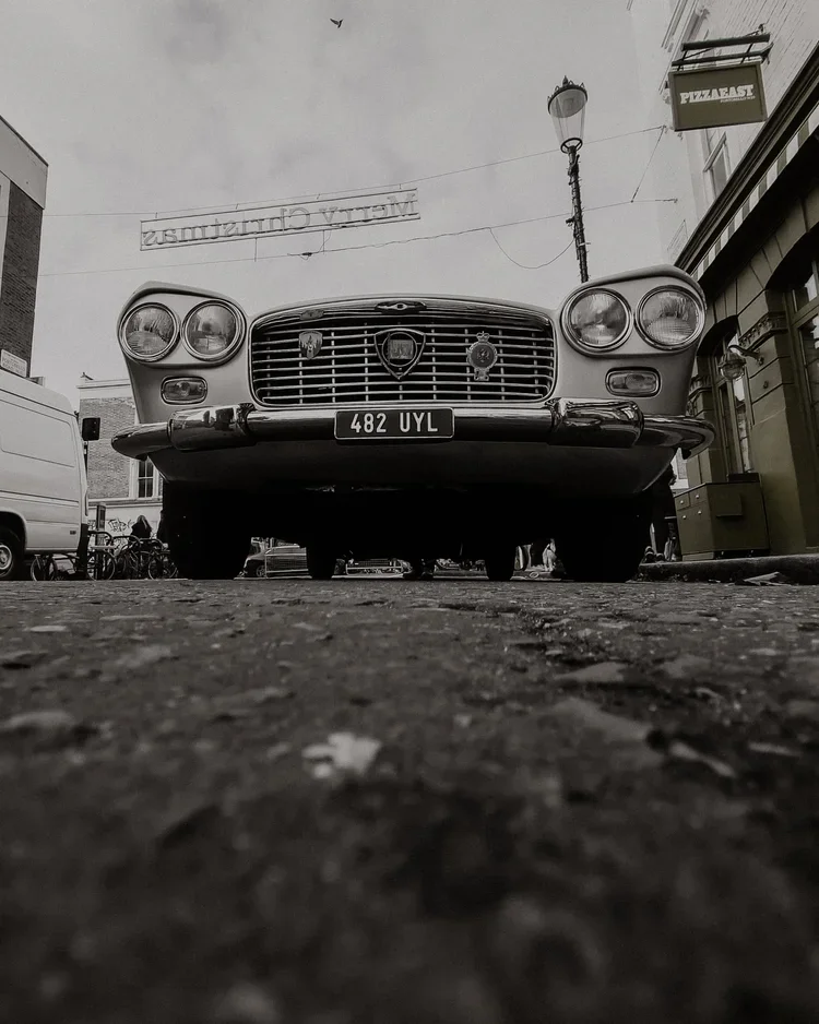 Black and white photograph of a vintage car parked on a cobblestone street from a low angle. The car has a European-style license plate reading '482 UYL' and is positioned in front of buildings with outdoor signage.